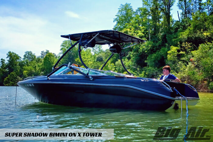 black boat sitting on the lake with lots of green trees around it. Showcasing the Big Air X tower and Super Shadow Bimini with a young man sitting in the back of the boat.