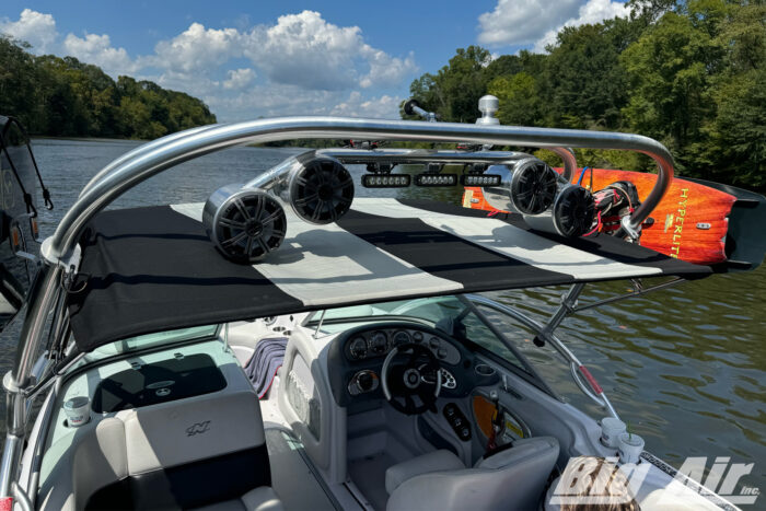 image showcases a speaker and light bar combo mounted onto a boat wake tower. Below the speaker and light bar combo is an open bimini on the boat that is black and white striped.