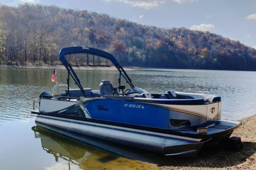 Blue 2023 Tahoe Tritoon boat beached on the water with a flag mounted on the rear pontoon tower