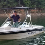 A boat on the water with a man at the helm. Boat has a wake tower and a bimini top