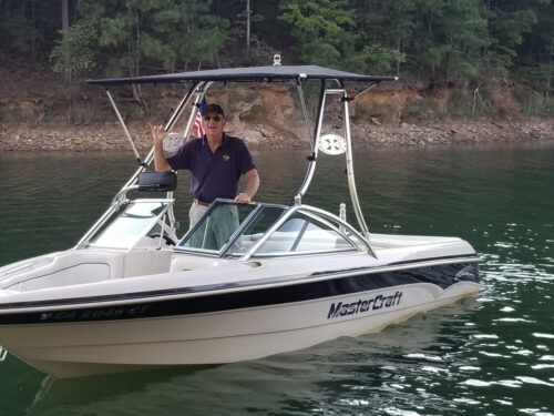 A boat on the water with a man at the helm. Boat has a wake tower and a bimini top